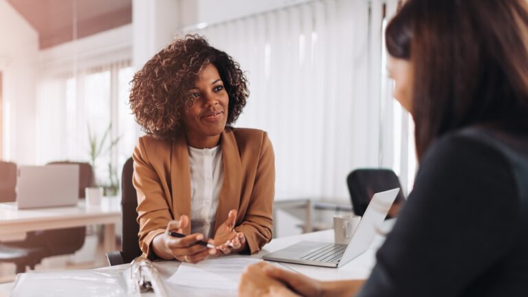 Artfully Media - Recruitment Advertising Made Simple Improve job advertising ROI without additional spend - Two ladies sat at a desk having a meeting. Happy looking using a laptop.